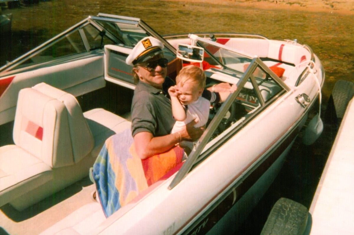 A 1990s photo of a grandfather in a captain's hat holding a toddler in a speedboat, before enhancement